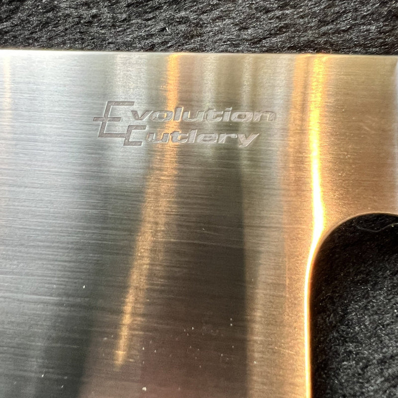 Close-up of a brushed stainless steel knife blade with the 'Evolution Cutlery' logo engraved on it, resting on a dark textured surface.