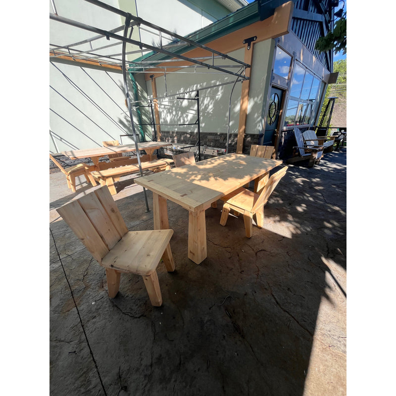 A white cedar dining table with heavy base legs sits on a patio of textured concrete, paired with matching dining chairs, set against the background of a green outdoor retailer's store.