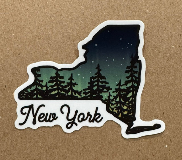 A die-cut sticker shaped like the state of New York, featuring a night sky with stars above a pine forest silhouette and the words "New York" in black script on a white background, placed on a brown textured surface.