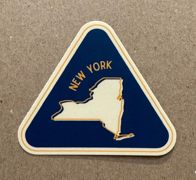 A triangular-shaped New York state  sticker with a dark blue background, cream outline, and gold text reading "NEW YORK," placed on a brown textured surface.