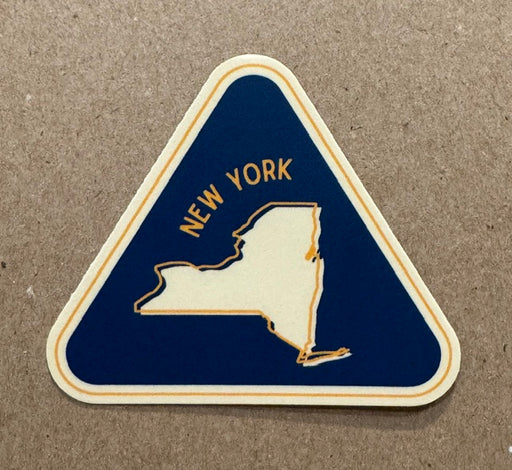 A triangular-shaped New York state  sticker with a dark blue background, cream outline, and gold text reading "NEW YORK," placed on a brown textured surface.