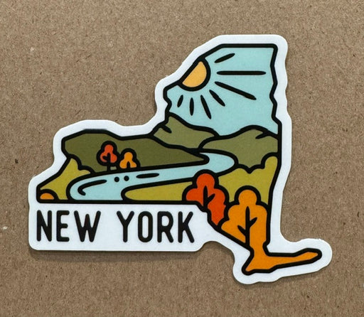 A die-cut sticker shaped like the state of New York, featuring a stylized landscape with a sun, mountains, trees, and a river, with the text 'NEW YORK' at the bottom, shown on a brown textured background.