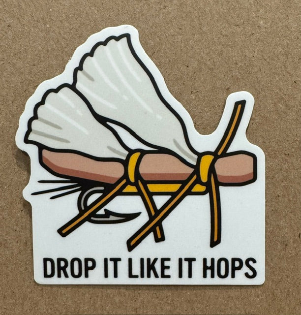 A die-cut sticker featuring a cartoon illustration of a fishing fly, with the text 'DROP IT LIKE IT HOPS' in black on a white background, placed on a brown textured surface.