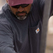 Man in a red hat and sunglasses wearing a carbon heather colored long sleeve shirt with a chest pocket. the chest pocket has a Carhartt logo on it. 