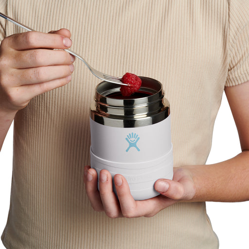 A person holding a white Hydro Flask insulated food container with a stainless steel lid, using a fork to put a raspberry into it. The person is wearing a beige ribbed short-sleeve top against a clean white background.