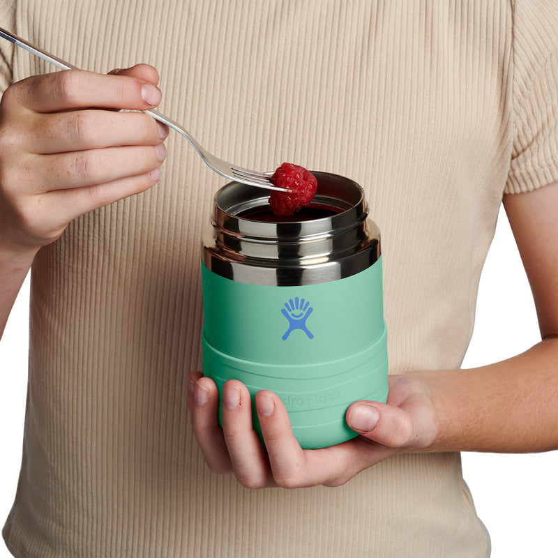 A person in a beige ribbed shirt holds a mint green Hydro Flask insulated container with a stainless steel lid, using a fork to add a red berry. The Hydro Flask logo is visible on the front.