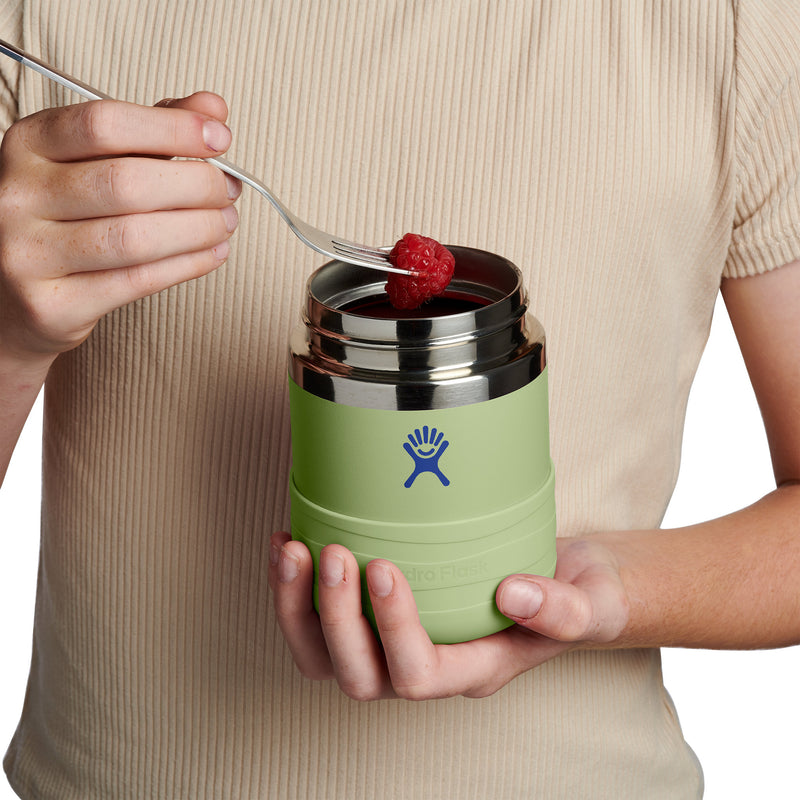 A person in a beige ribbed shirt holds a light green Hydro Flask insulated food jar with a stainless steel lid, using a fork to put a raspberry into it, against a white background.