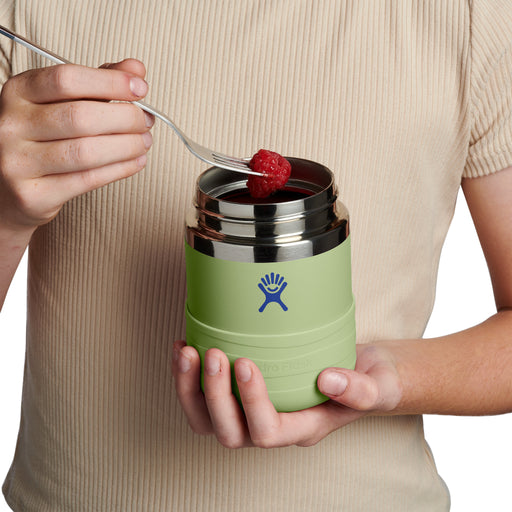 A person in a beige ribbed shirt holds a light green Hydro Flask insulated food jar with a stainless steel lid, using a fork to put a raspberry into it, against a white background.
