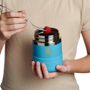 A person in a beige ribbed shirt holds a bright blue insulated container with a stainless steel lid while using a spoon to add a red berry. The container has a yellow logo on the sleeve.