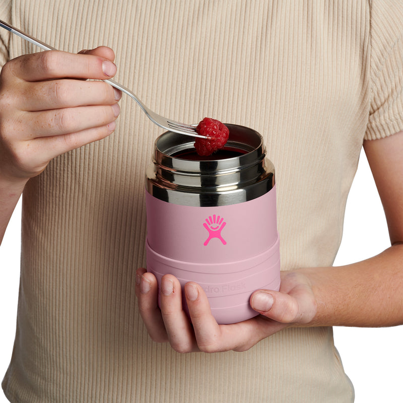 A person in a beige ribbed top holds a pink Hydro Flask insulated container with a stainless steel lid while feeding a raspberry onto a fork.