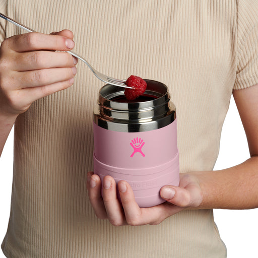 A person in a beige ribbed top holds a pink Hydro Flask insulated container with a stainless steel lid while feeding a raspberry onto a fork.