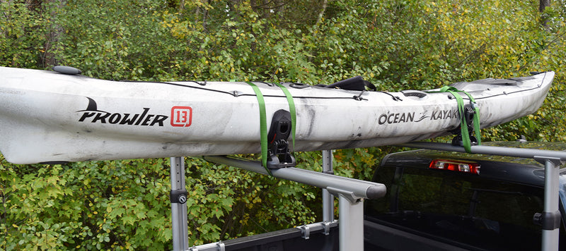 Side view of a white kayak strapped to a truck against some foliage. 