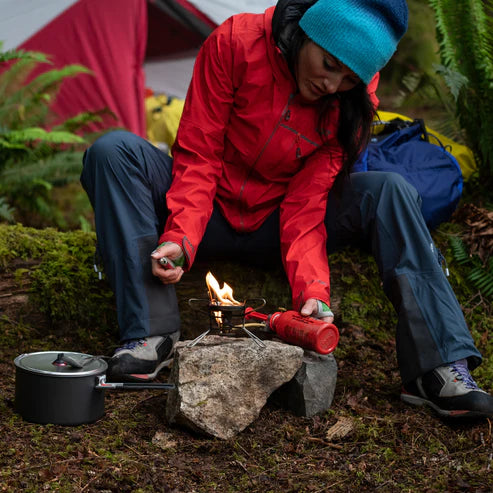 Person in red jacket and blue beanie preparing a campfire with a pot nearby, camping setting.