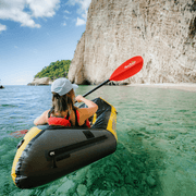 Person paddling a yellow inflatable kayak through clear water with a rocky cliff in the background.