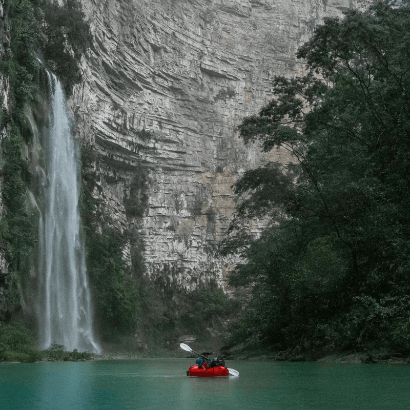 Kayaker in a red kayak paddling towards a waterfall in a lush green forest.