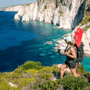 Person with red paddles in their backpack standing on a cliff overlooking the ocean with a rocky cliff in the background.