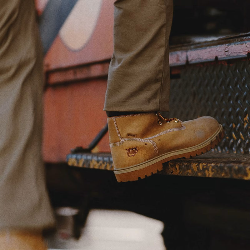 A close-up of a person's foot wearing a tan lace-up work boot, stepping onto a rusty metal platform next to a red vehicle.