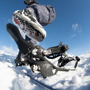 A person stepping into a black snowboard binding with a Nidekcer logo on the back on a snowy hill. 