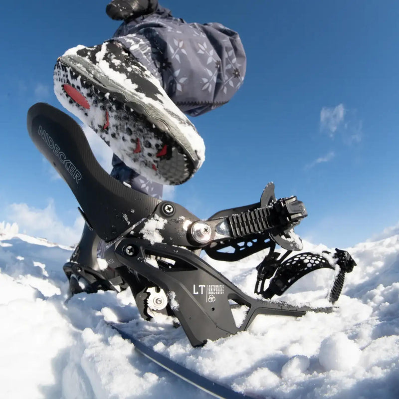 A person stepping into a black snowboard binding with a Nidekcer logo on the back on a snowy hill. 