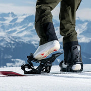A person putting their foot into a black snowboard binding on a snowy mountain.