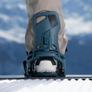 Person standing on a snowboard with their foot in a blue colored snowboard binding on a snowy hill. 