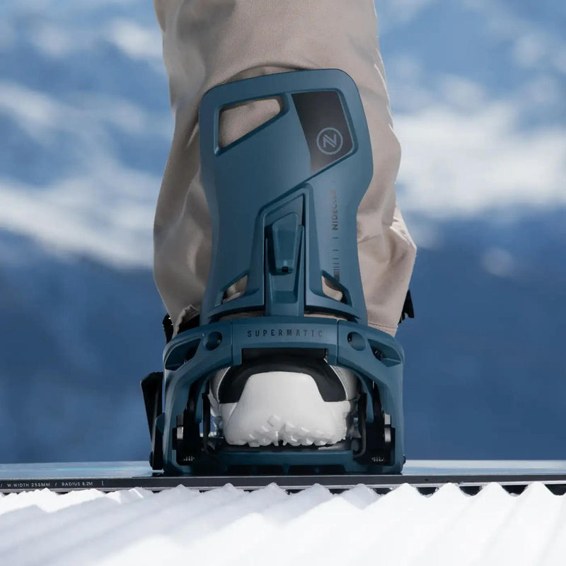 Person standing on a snowboard with their foot in a blue colored snowboard binding on a snowy hill. 