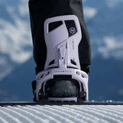A person standing on a snowboard with their foot in a white binding with a Nidecker logo on a snowy hill. 