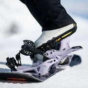 A person putting their foot into a white snowboard binding with black accents on a snowy mountain.