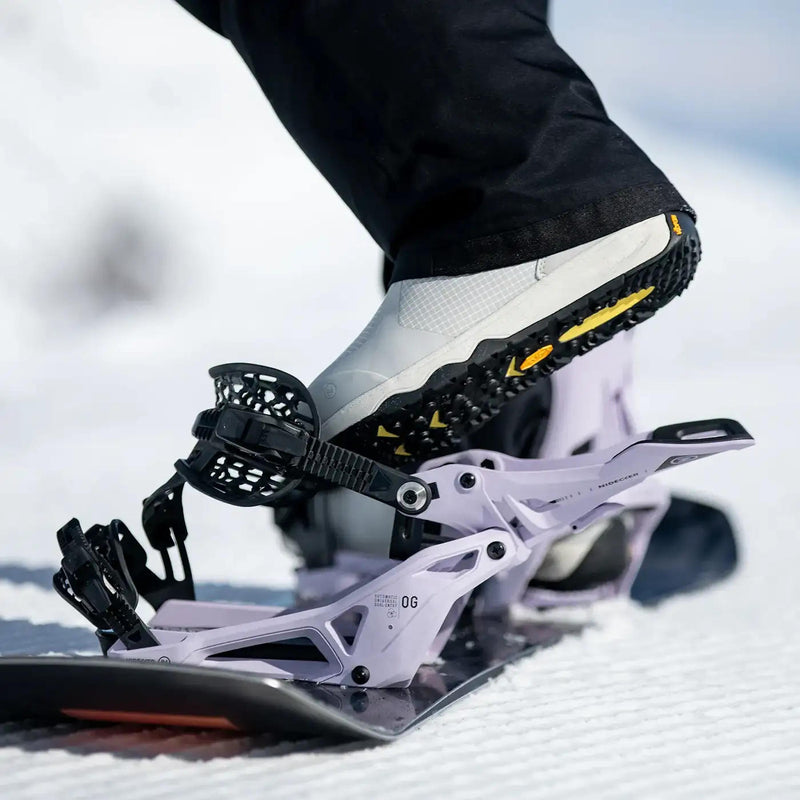 A person putting their foot into a white snowboard binding with black accents on a snowy mountain.