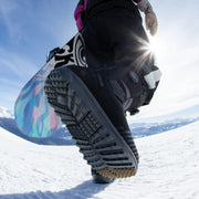 A person carrying a snowboard wearing a pair of black snow boots with a Nidecker label on a snowy mountain. 
