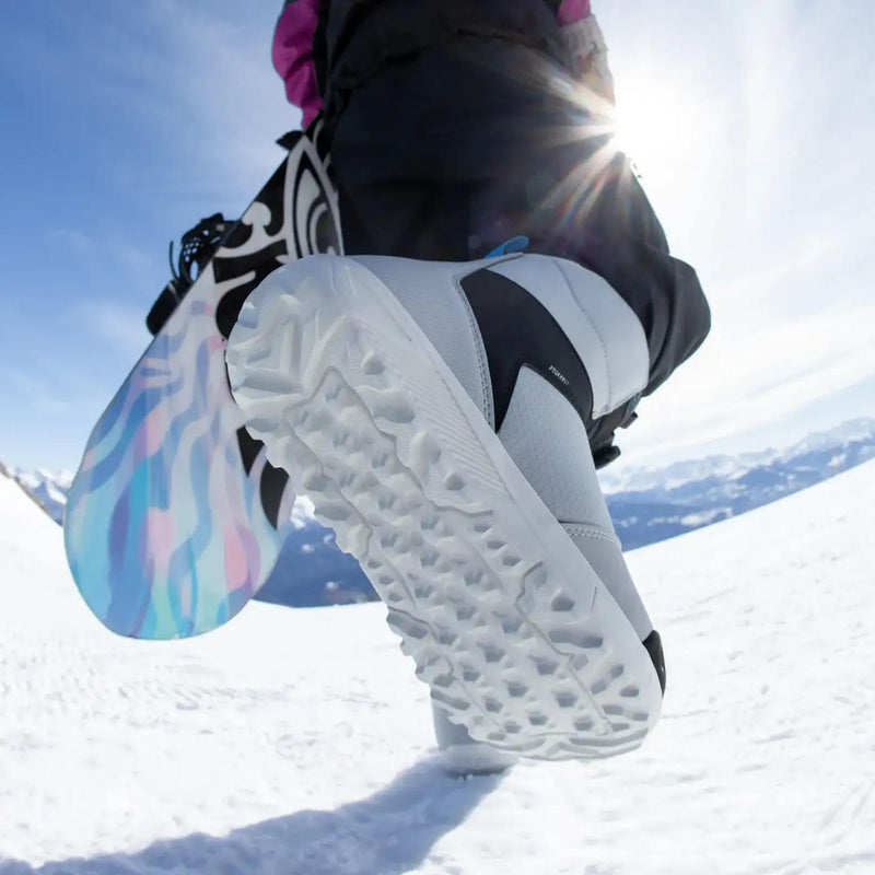 A person holding a snowboard walking on a snowy mountain wearing white snow boots.