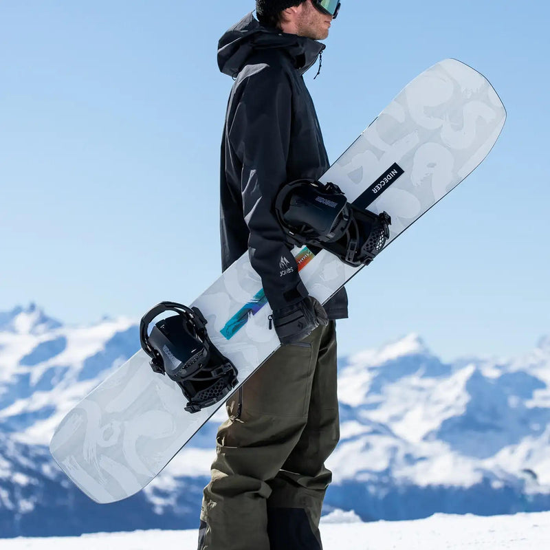 Side view of a man holding a white snowboard with a Nidecker logo on a snowy hill with mountains in the background. 
