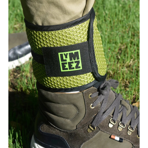 A lime green and black ankle wrap worn around a person's ankle, paired with brown hiking boots and khaki pants, set against a grassy outdoor background. The wrap features a black strap with the brand name 'LYMEEZ' in neon green.