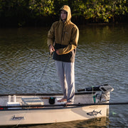 A man in a brown jacket standing on a white boat holding a fishing rod with water in the background.