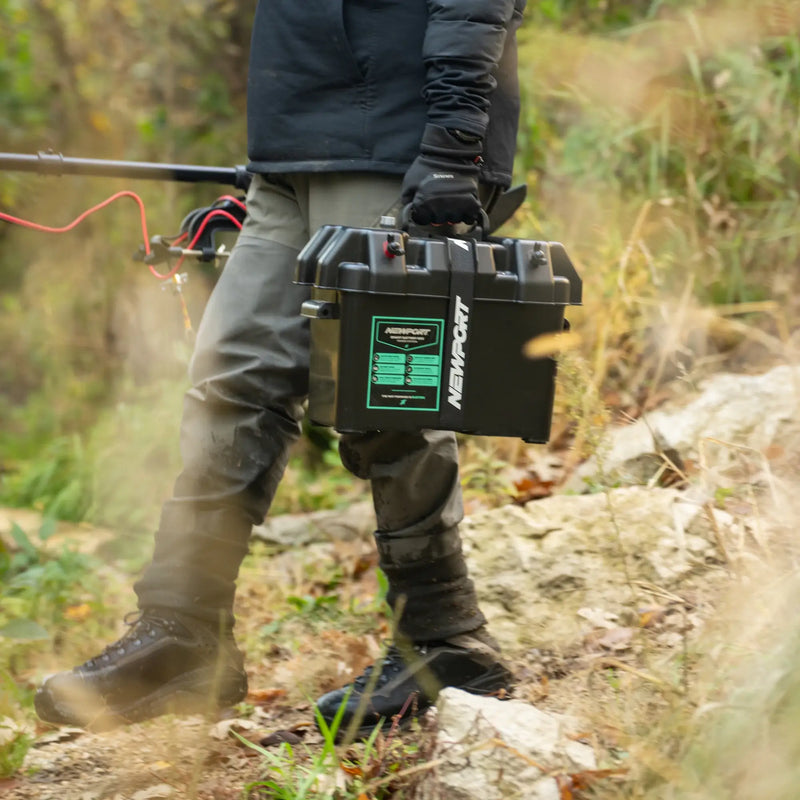 A person carrying a battery box through a forest with a visible Newport logo on it.
