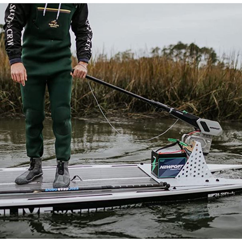 A person stands on a paddleboard on a body of water, holding a long handle extension for the tiller on his motor. The board has a battery mounted on it, and the background shows reeds and water.