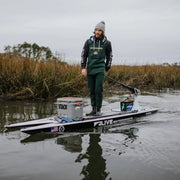 A man stands on a paddleboard rigged for fishing, using a tiller handle extension to steer the motor mounted to the back