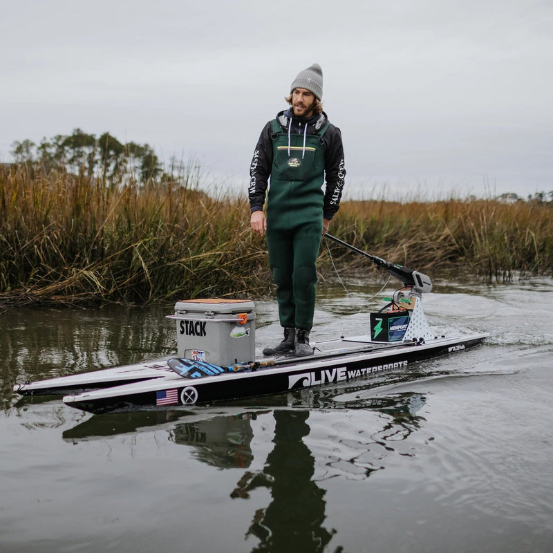 A man stands on a paddleboard rigged for fishing, using a tiller handle extension to steer the motor mounted to the back
