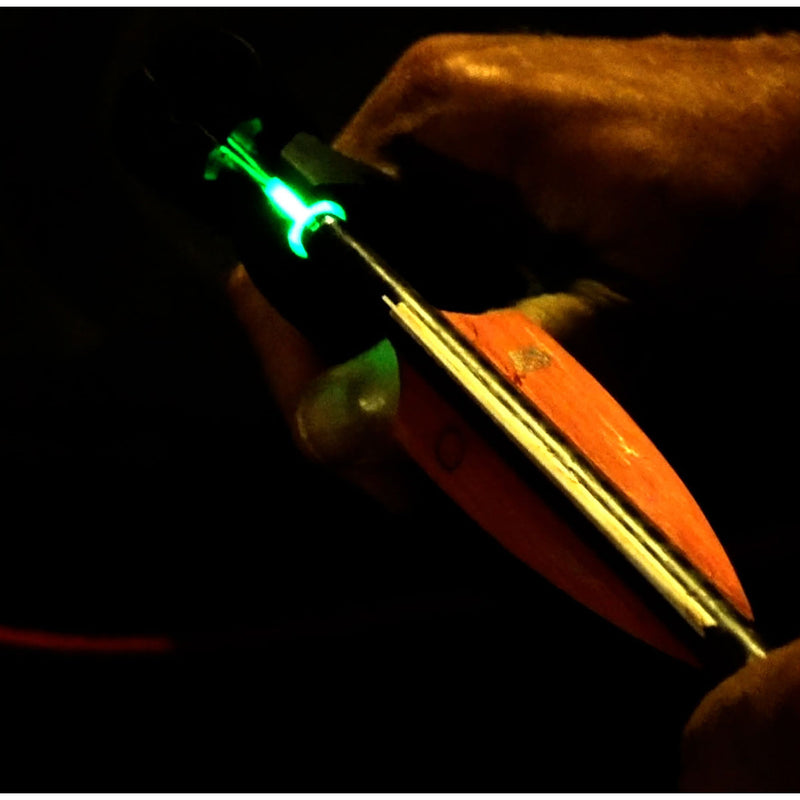 A close-up of a hand holding a bright green illuminated nock and nock-deactivating tool against a very dark background.