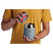A person in a mustard yellow t-shirt holds a light blue insulated bottle with a stainless steel lid, showing a red seal and the blue silicone seal being lifted by the other hand.