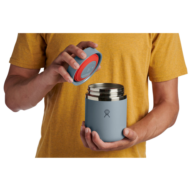 A person in a mustard yellow t-shirt holds a light blue insulated bottle with a stainless steel lid, showing a red seal and the blue silicone seal being lifted by the other hand.