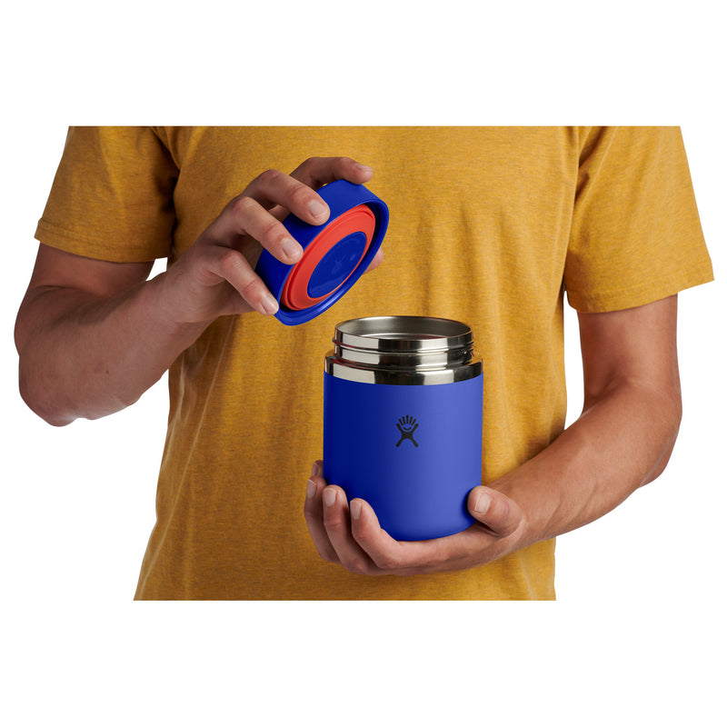 A person in a mustard yellow t-shirt holds a blue insulated travel tumbler with a stainless steel interior and a blue lid with a red seal, against a plain white background.