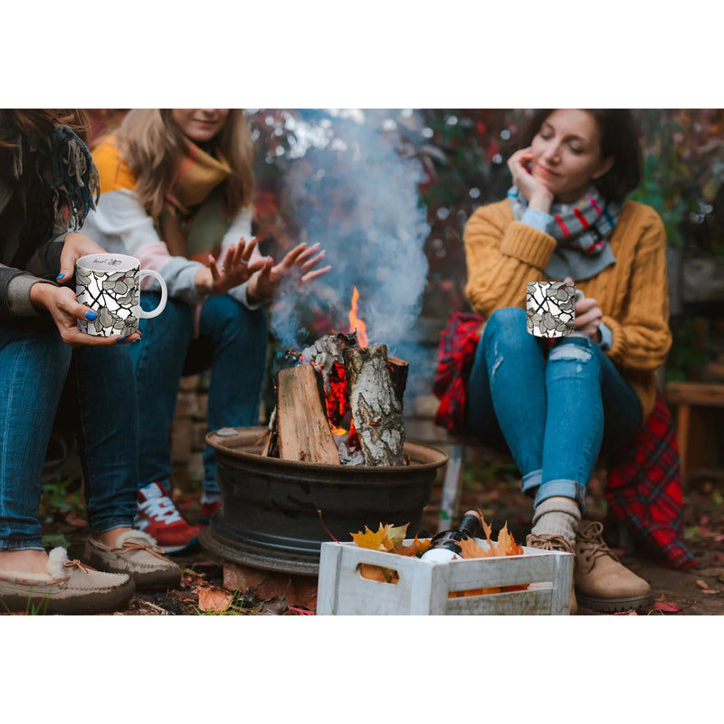 Three women in cozy sweaters and flannels sit around a small fire pit. Two of them are holding coffee mugs printed with Tiffany's Magnolias stained glass design.