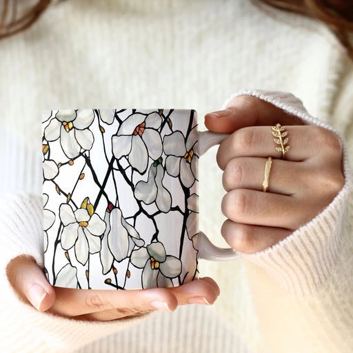A person in a cream ribbed sweater holds a white ceramic mug decorated with a stained-glass style floral pattern of white blossoms and black outlines. The person is wearing two gold rings on their fingers.