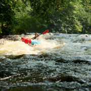 Woman in a red and blue kayak paddling through rough waters with trees in the background.