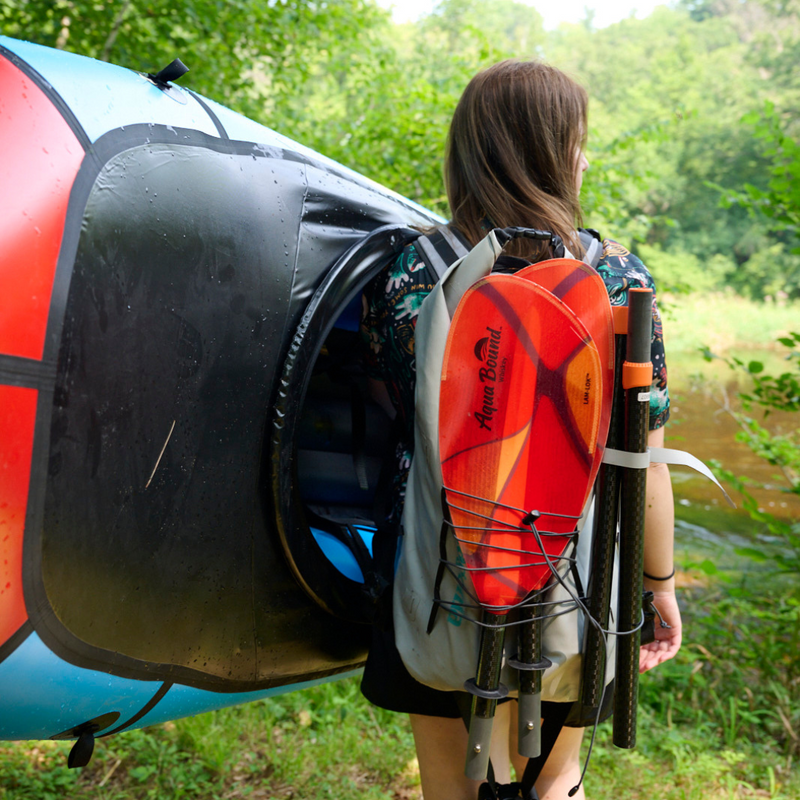 Person carrying a kayak and paddle on a trail near a body of water