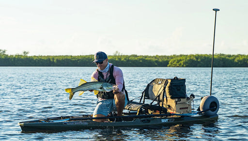 Man holding a fish in a dark green fishing kayak on a lake with trees in the background.