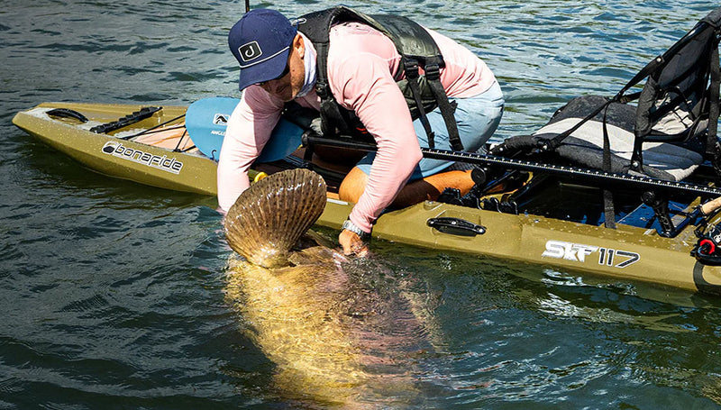 Man wrangling a big fish on a green fishing kayak in lake.
