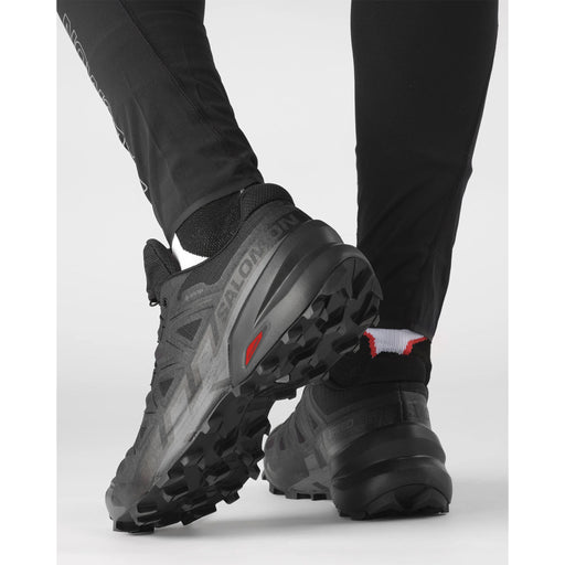 A pair of black-on-black trail running sneakers with heavy grip tread outsoles, shown from the rear on a model