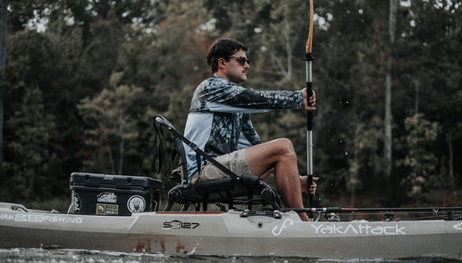 Man paddling his grey kayak in a lake with trees in the background.